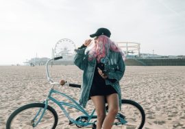 Woman with pink hair posing near a bicycle on the beach.