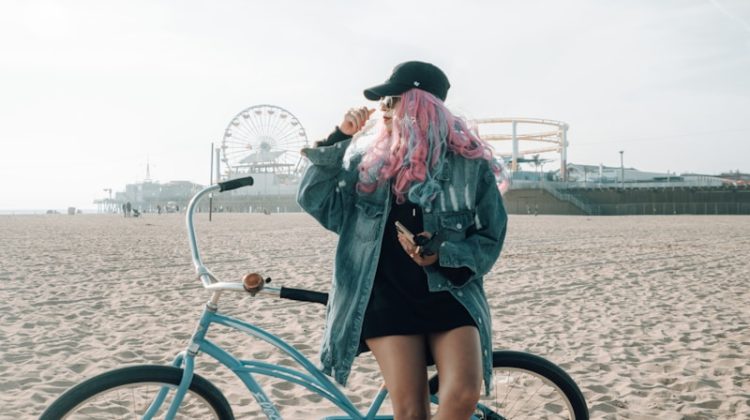 Woman with pink hair posing near a bicycle on the beach.
